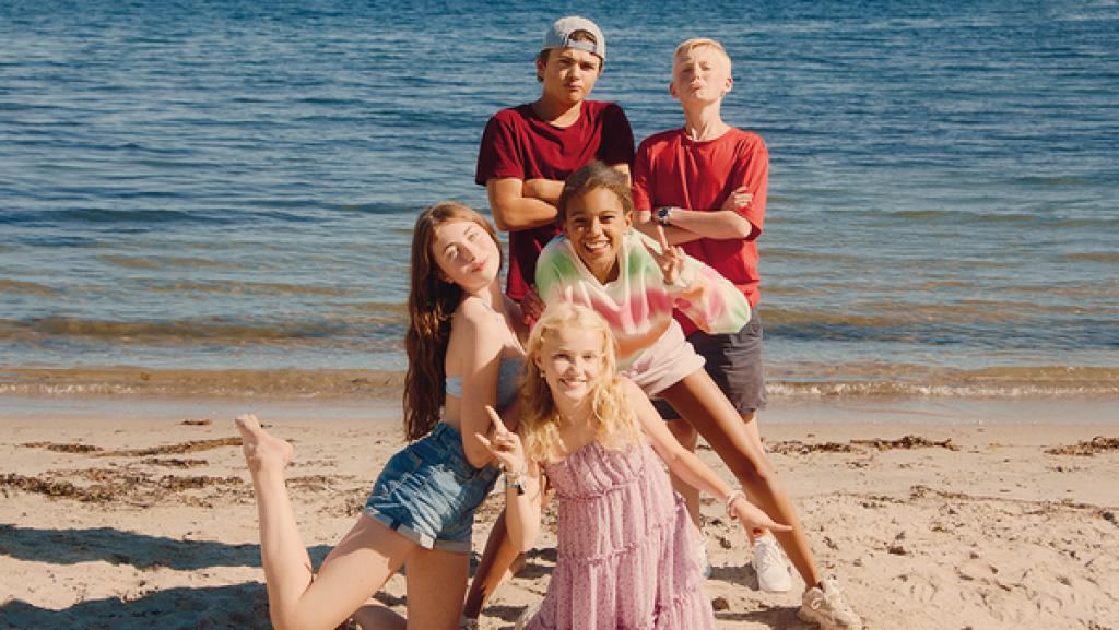 5 Jugendliche posieren am Strand, dahinter das Meer. Zwei Jungen stehen hinter drei Mädchen.